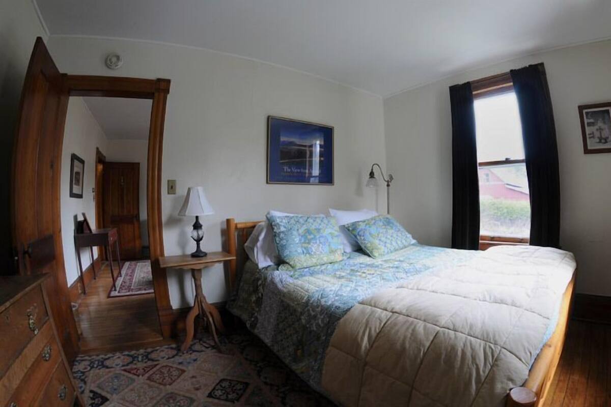 A bedroom with a bed, a desk, a chair, a lamp, and a door leading to another room.
