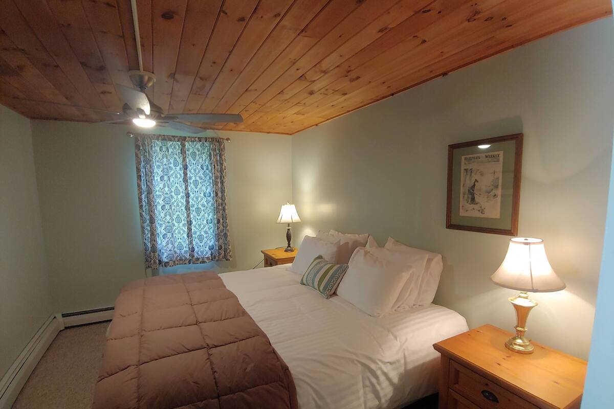 A bedroom with a wooden ceiling, a bed with a brown quilt, two bedside tables with lamps, a window with lace curtains, and a framed picture on the wall.