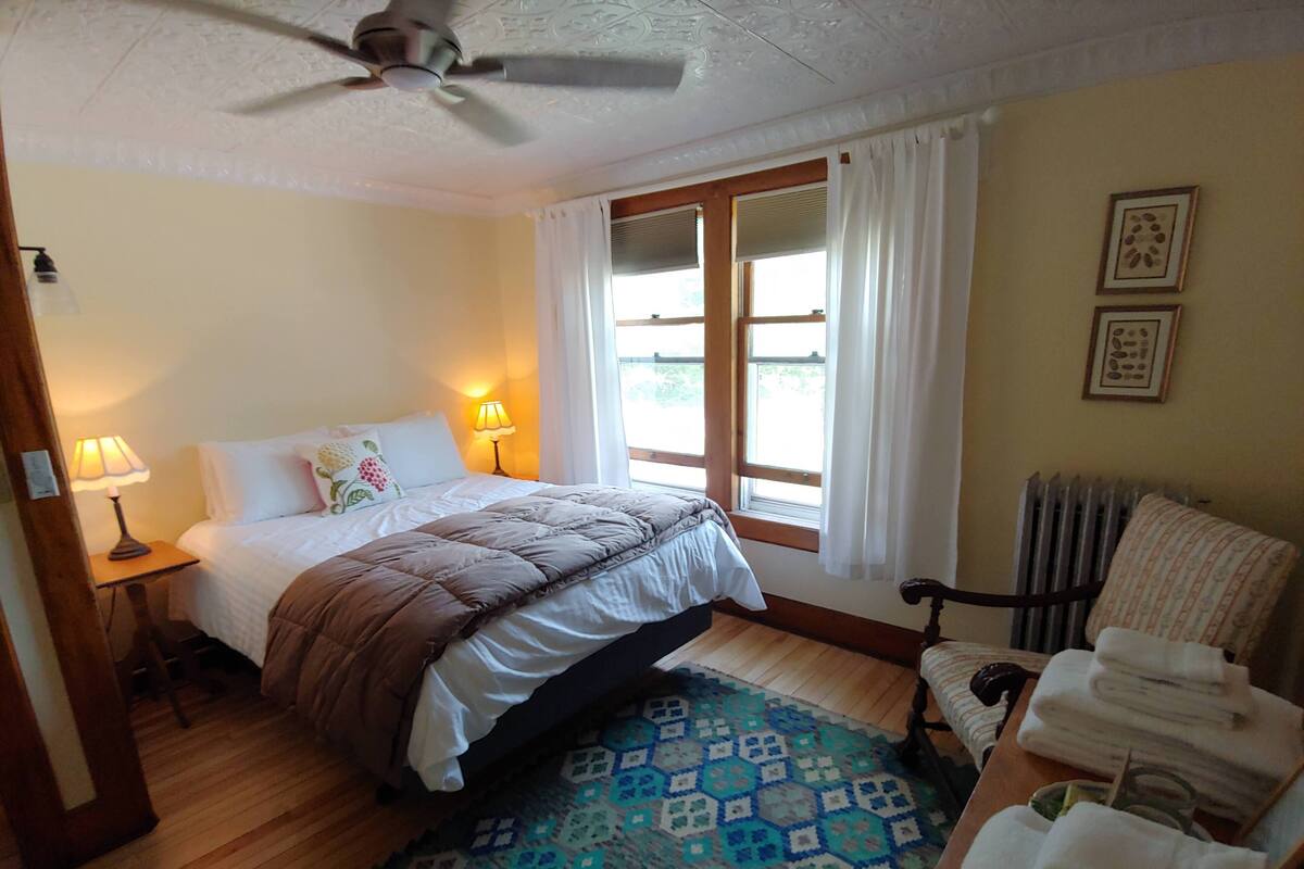 A bedroom with a bed, bedside lamps, a patterned rug, a chair, and a radiator.
