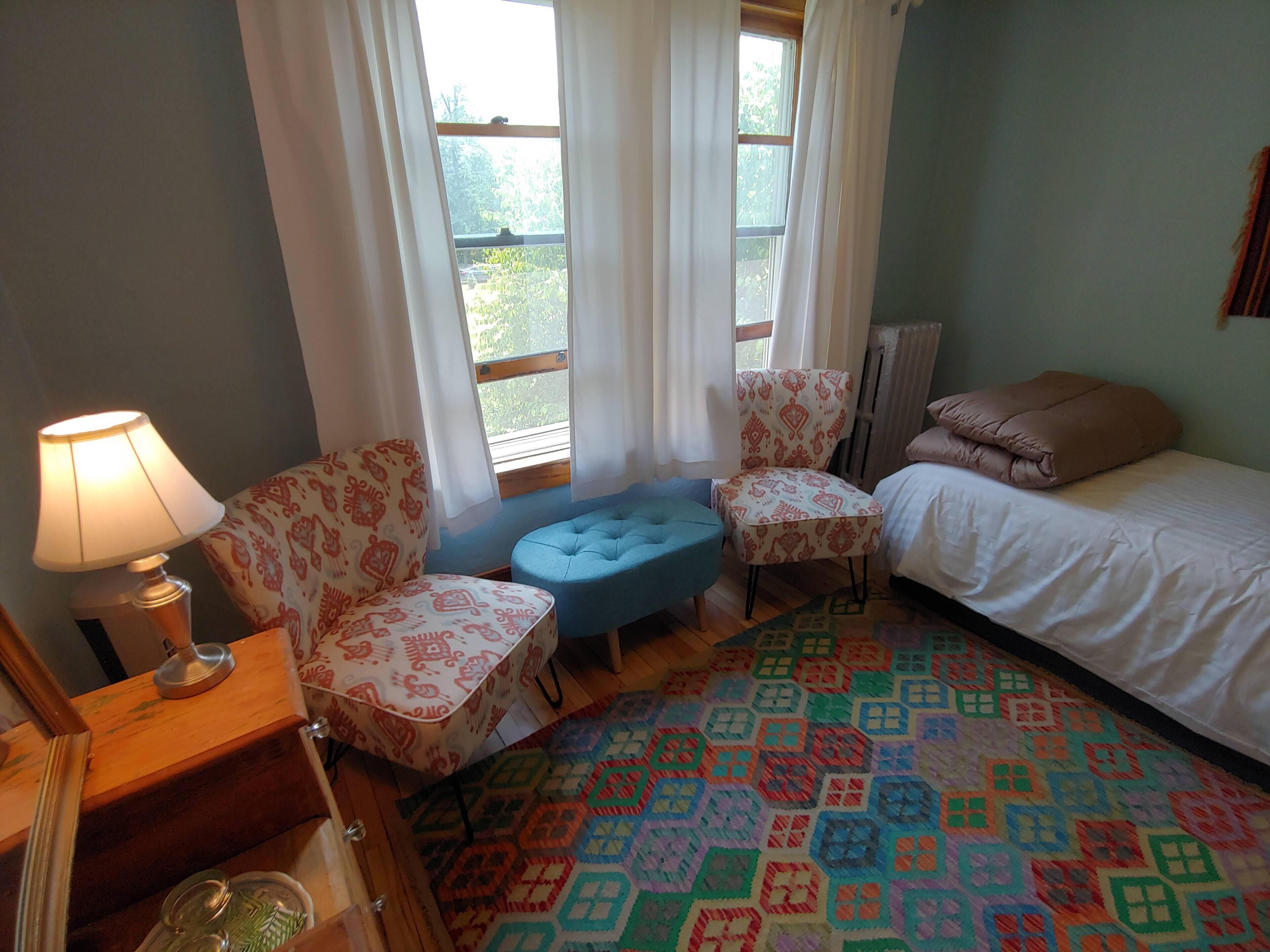 A room with a bed, two patterned chairs, a small blue ottoman, a lamp, and a colorful patterned rug.