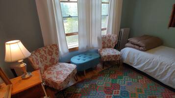 A room with a bed, two patterned chairs, a small blue ottoman, a lamp, and a colorful patterned rug.