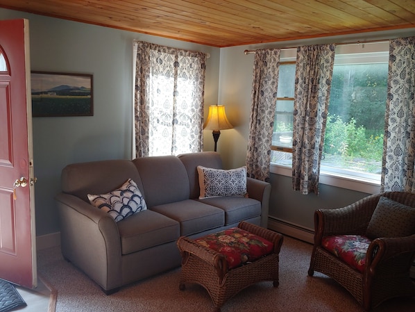 A cozy living room with a sofa, a wicker ottoman, and a chair. There is a lamp on a side table, and curtains on the windows. - Keene Valley Lodge (Keene Valley)
