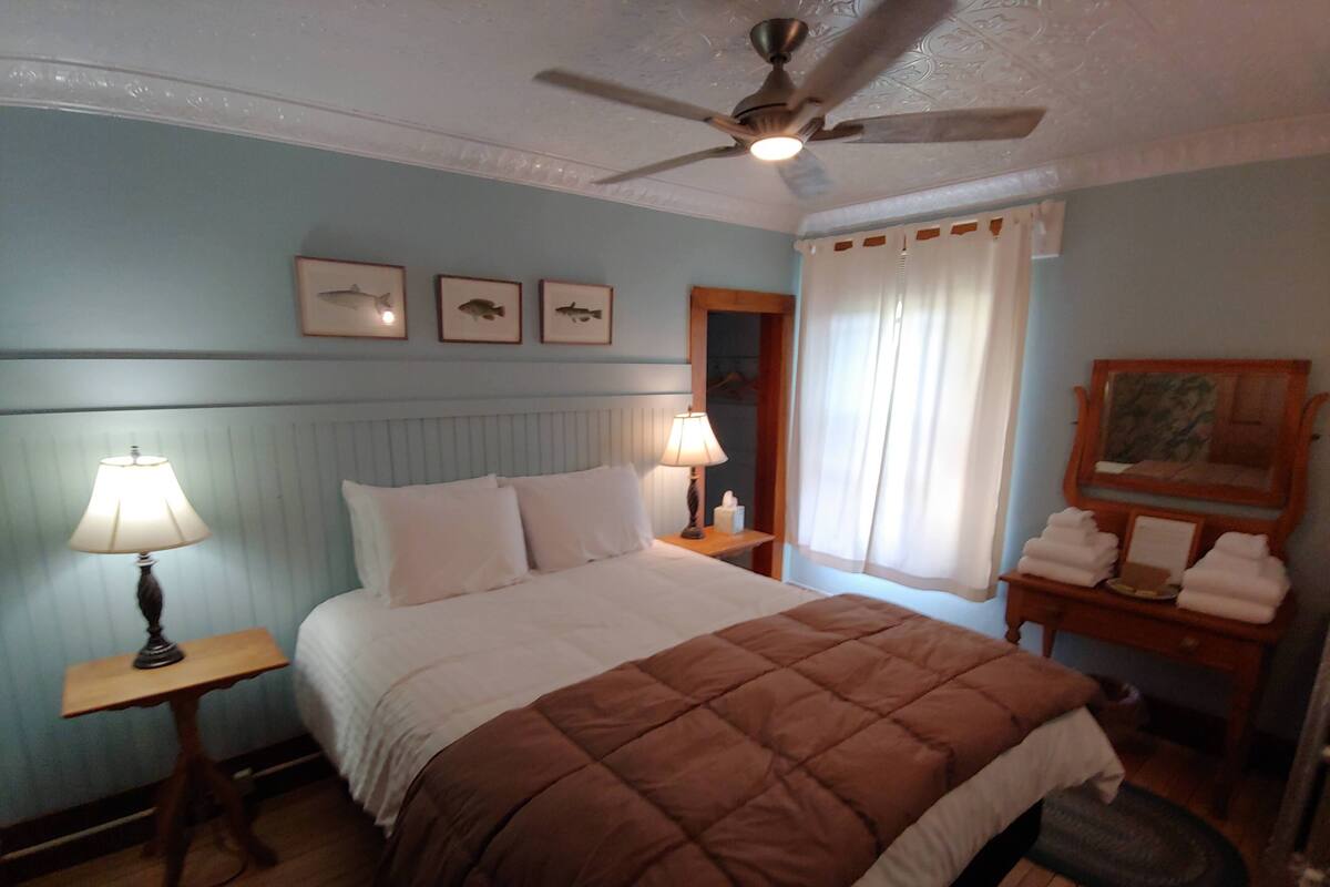 A bedroom with a bed, a nightstand, a lamp, a ceiling fan, a window with curtains, and a wall with framed pictures.