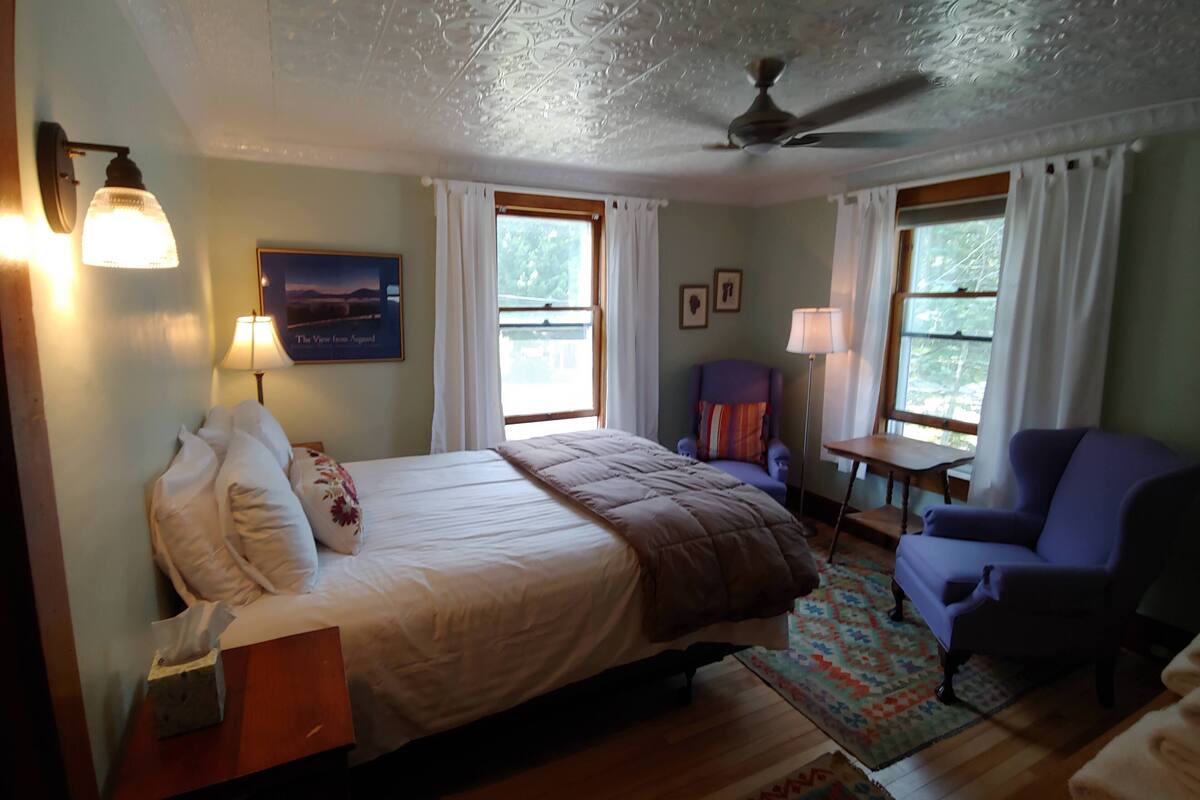 A bedroom with a bed, two chairs, a nightstand, a lamp, and a ceiling fan.