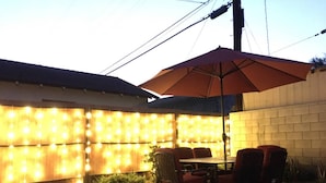 Un patio exterior con una mesa y sillas, un paraguas grande y luces colgantes en una pared.