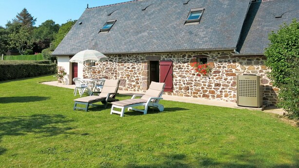 A stone house with a sloped roof, a chimney, and a small window. There are two lounge chairs and a table in the front yard.