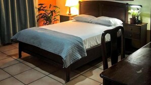 A neatly made bed with a blue quilt, a bedside lamp, a small table with a plant, and a wall-mounted vase. - Hotel Alsacia (Tegucigalpa)