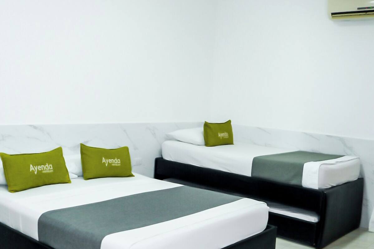 Een moderne hotelkamer met twee bedden, elk voorzien van een groen kussen met de tekst 'Ayenda', een witte muur en een aan het plafond bevestigde airconditioner.