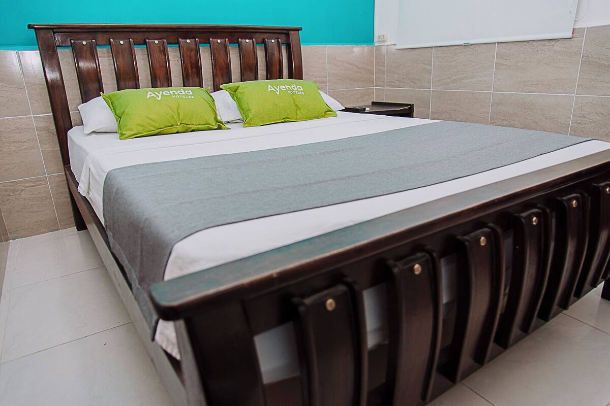 Een bed met houten frame, groene kussens en een grijze spreidek, in een kamer met betegelde muren.