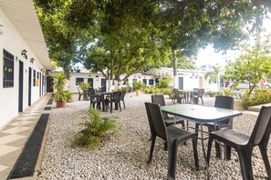 Un espace extérieur aménagé en terrasse, avec des tables et des chaises, entouré de bâtiments blancs et de verdure.