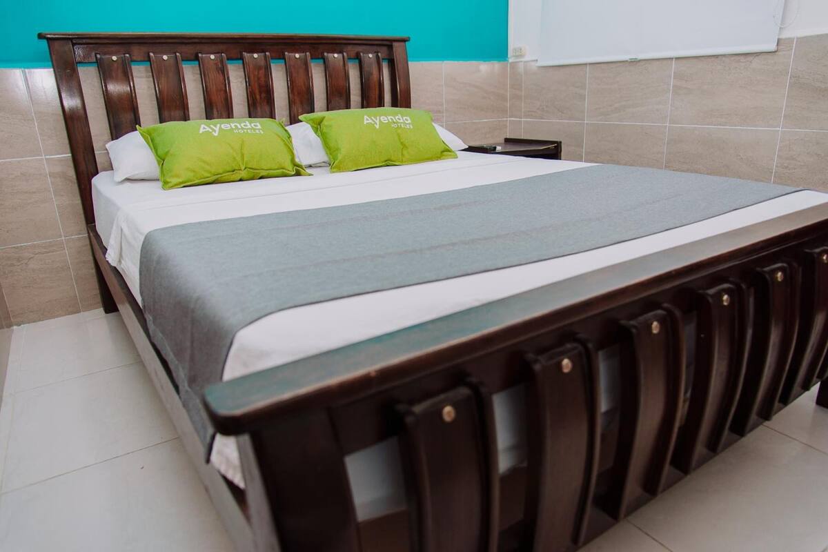 Een bed met een houten hoofdbord, twee groene kussens en een grijze deken.