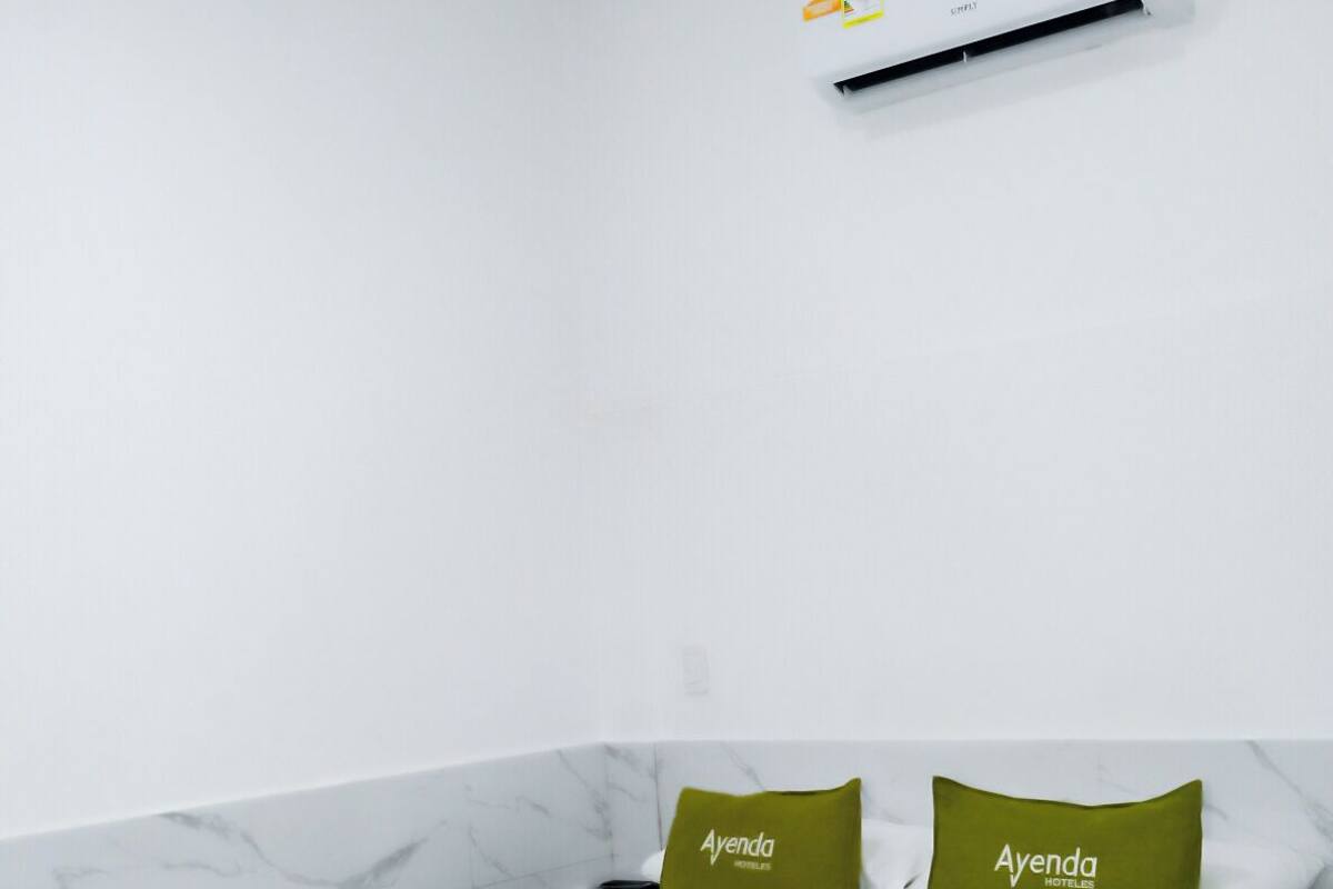 Een hotelkamer met een bed, twee kussens met 'Ayenda Hotel'-logo, een witte airconditioningunit aan de muur en een wand met marmerpatroon.