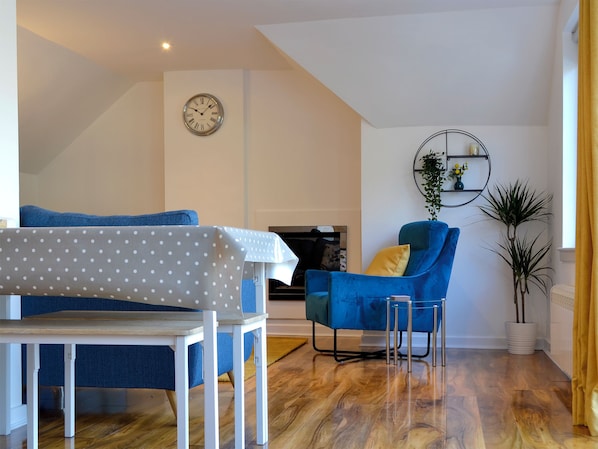 A modern living room with a blue armchair, a small table with a polka dot tablecloth, a clock on the wall, and a potted plant.