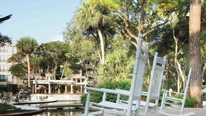 Ein Uferbereich mit Restaurant, Pool und weißen Schaukelstühlen am Wasser.