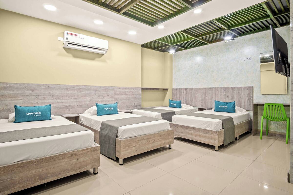 Habitación de hotel con cuatro camas individuales, cada una con cabecero gris y almohada azul con la etiqueta 'ayenda'. Hay una silla verde, un televisor de pantalla plana montado en la pared y un equipo de aire acondicionado sobre las camas.