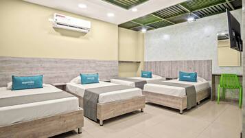 Quarto de hotel com quatro camas de solteiro, cada uma com cabeceira cinza e travesseiro azul com a inscrição 'ayenda'. Há uma cadeira verde, uma televisão de tela plana presa à parede e um ar-condicionado sobre as camas.