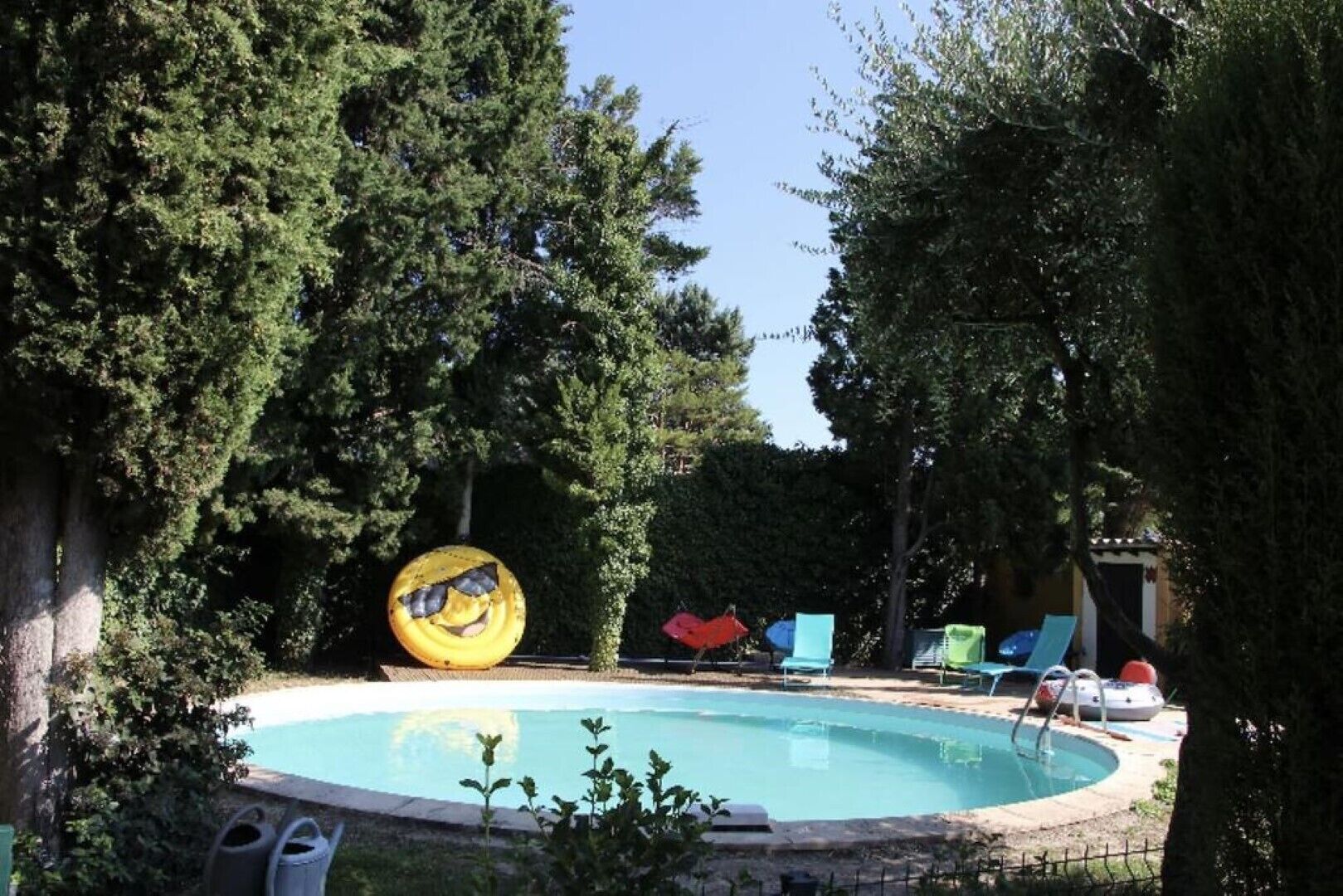 Una piscina rodeada de árboles, con una pelota amarilla grande y varias sillas de descanso.