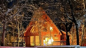 A wooden cabin with a lit interior, surrounded by snow-covered trees. - Charming Petersen Cottage Rustic Luxury  (Pine Valley)