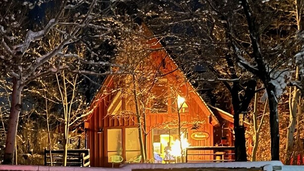 Une cabane en bois, dont l’intérieur est éclairé, entourée d’arbres couverts de neige.