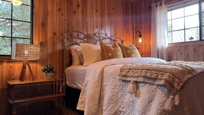 A wooden-paneled bedroom with a bed, a bedside table with a lamp, and a window with curtains. - Charming Petersen Cottage Rustic Luxury  (Pine Valley)