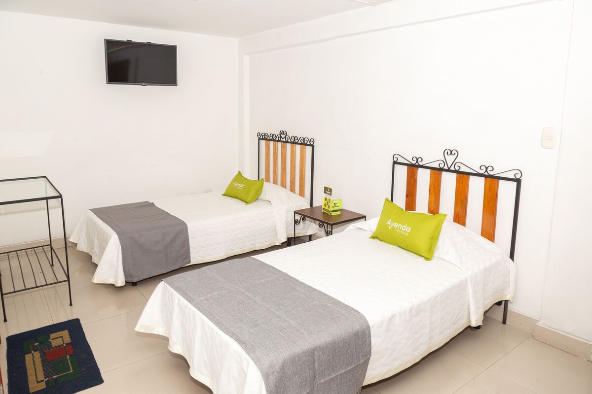 Une chambre d’hôtel avec deux lits simples, un téléviseur fixé au mur et une petite table sur laquelle se trouve une boîte.
