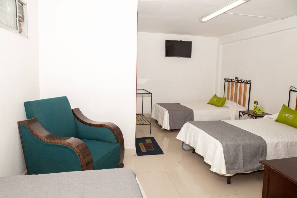 A hotel room with two single beds, a teal armchair, a TV on the wall, and a small table with a green pillow.