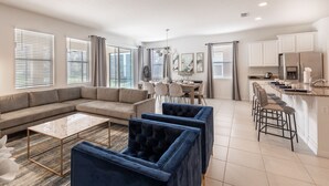 A modern living room with a sectional sofa, a coffee table, and two armchairs. There is a dining area with a table and chairs, and a kitchen with a refrigerator and bar stools. - 5268OBA: Outdoor Projector 6bed in Solterra minutes from major Orlando attractions (Davenport)