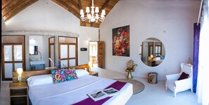 A spacious bedroom with a large bed, a chandelier, a mirror, and a view of the outside.