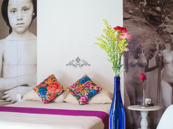 A hotel room with a bed, colorful pillows, a vase with flowers, a small table with a lamp, and a large wall mural.