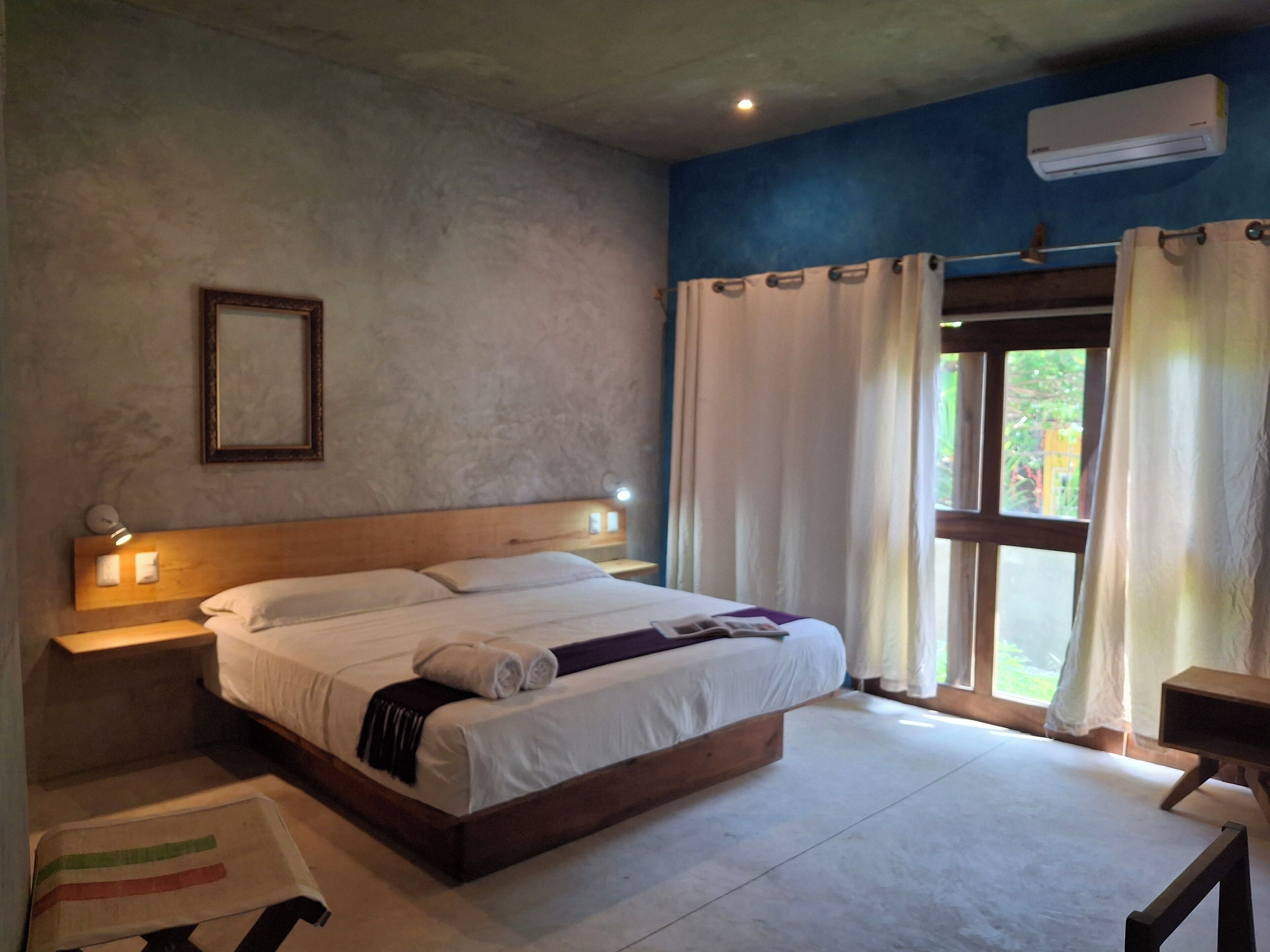A bedroom with a large bed, bedside lamps, a wooden headboard, a nightstand, a window with curtains, and an air conditioning unit.