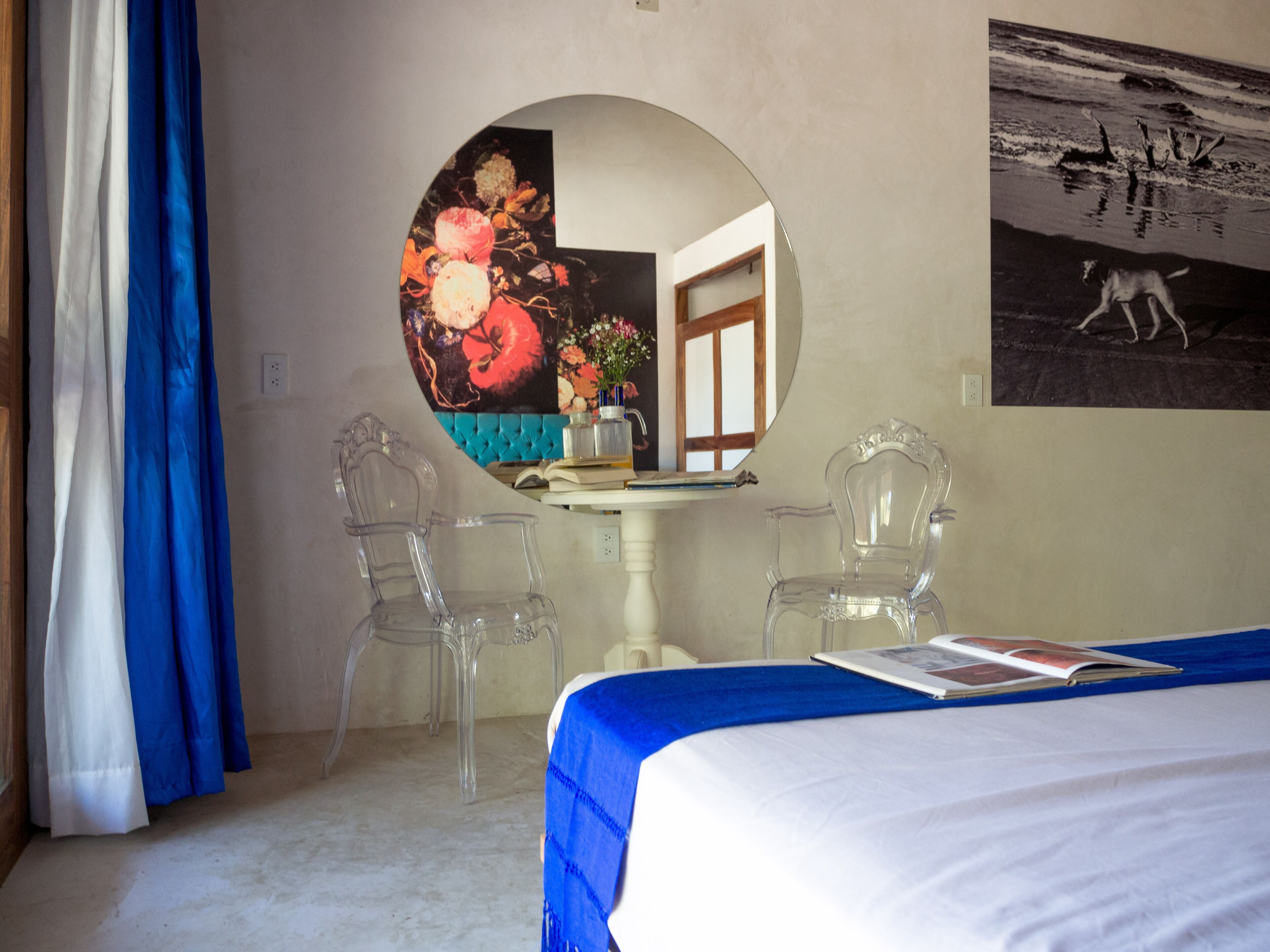 Una habitación de hotel con una cama, dos sillas traslúcidas, un espejo redondo, un cuadro y una ventana.
