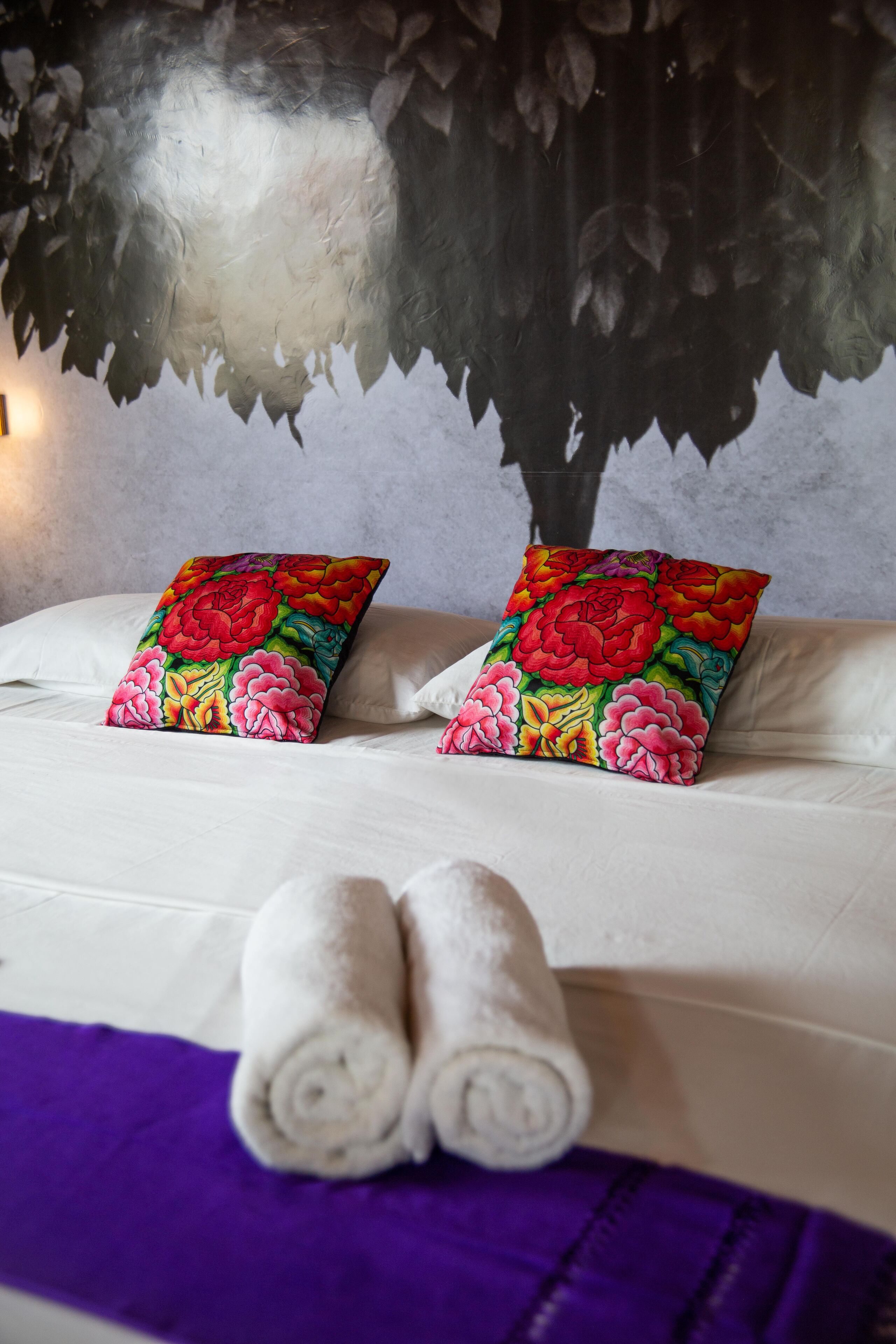 Una cama con dos almohadas de estampado floral, una toalla enrollada y un edredón morado.