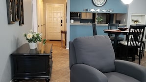 A living room with a grey armchair, a dark wooden coffee table with a vase of flowers, and a kitchen in the background with a blue wall and a clock. - Hidden Treasure! Charming ground floor condo in waterfront complex. (Marble Falls)