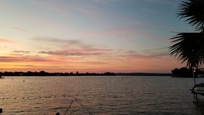 A calm body of water with a sunset sky, a palm tree, and a boat docked on the right.