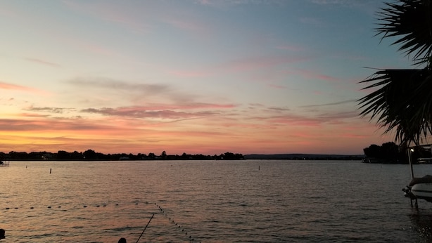 A calm body of water with a sunset sky, a palm tree, and a boat docked on the right. - Hidden Treasure! Charming ground floor condo in waterfront complex. (Marble Falls)