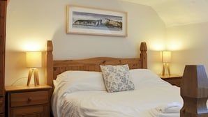 Un lit bien fait, avec du linge de lit blanc et un coussin à motifs floraux, flanqué de deux tables de chevet en bois surmontées de lampes, dans une pièce où un tableau représentant un paysage côtier encadré orne le mur.