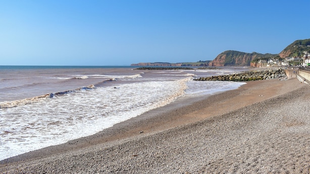 Une plage côtière avec des vagues, un rivage rocheux et des bâtiments en arrière-plan.