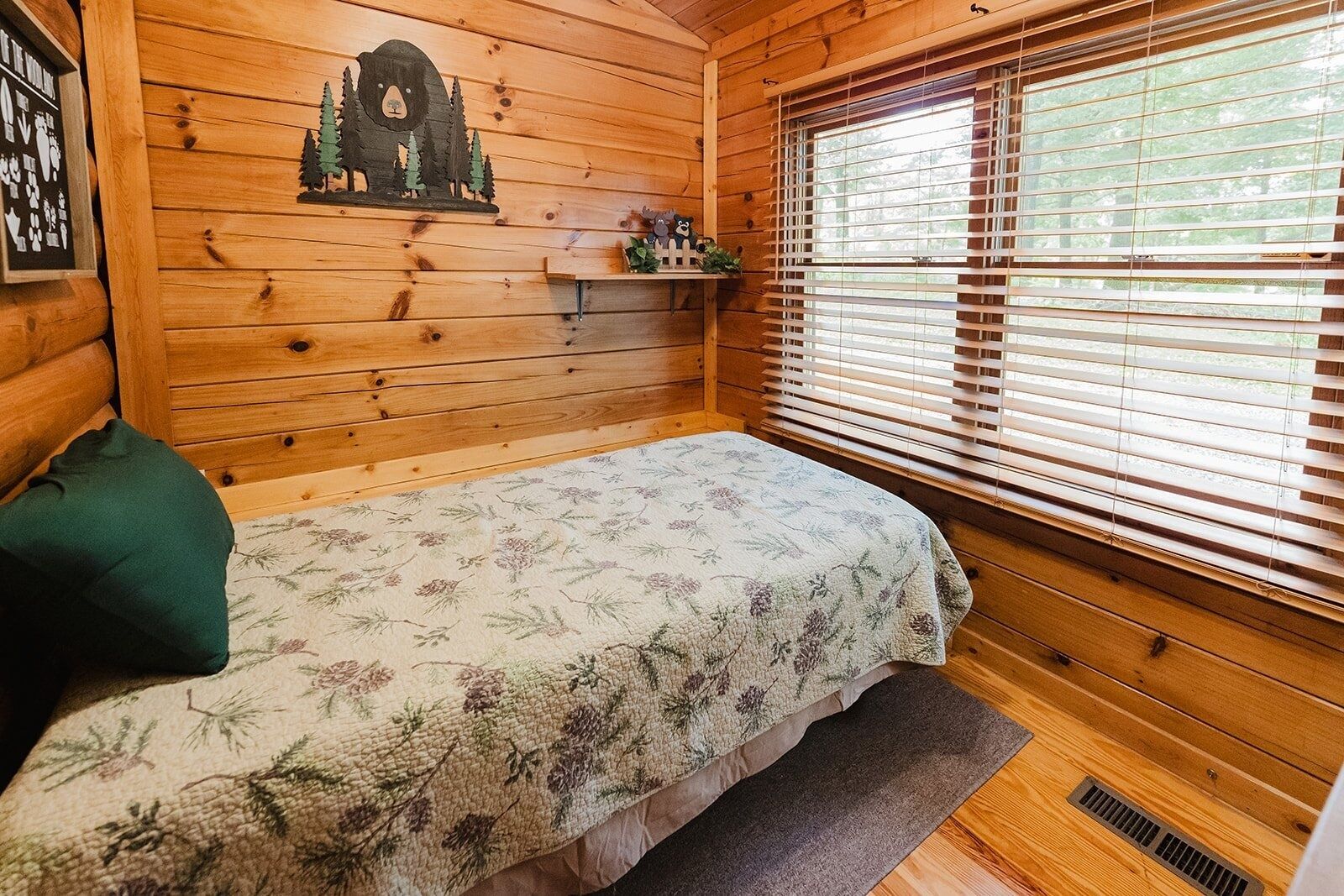 Una habitación de cabaña acogedora con una cama, una pared de madera con un recorte de oso, una ventana con persianas y un estante pequeño con una planta.