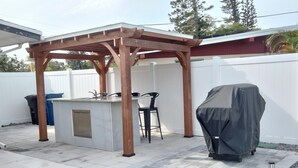 A covered outdoor kitchen area with a pergola structure, a bar counter, and two bar stools. - Beach Style House w/ large backyard, close to fishing, canoeing, and beaches (Bradenton)