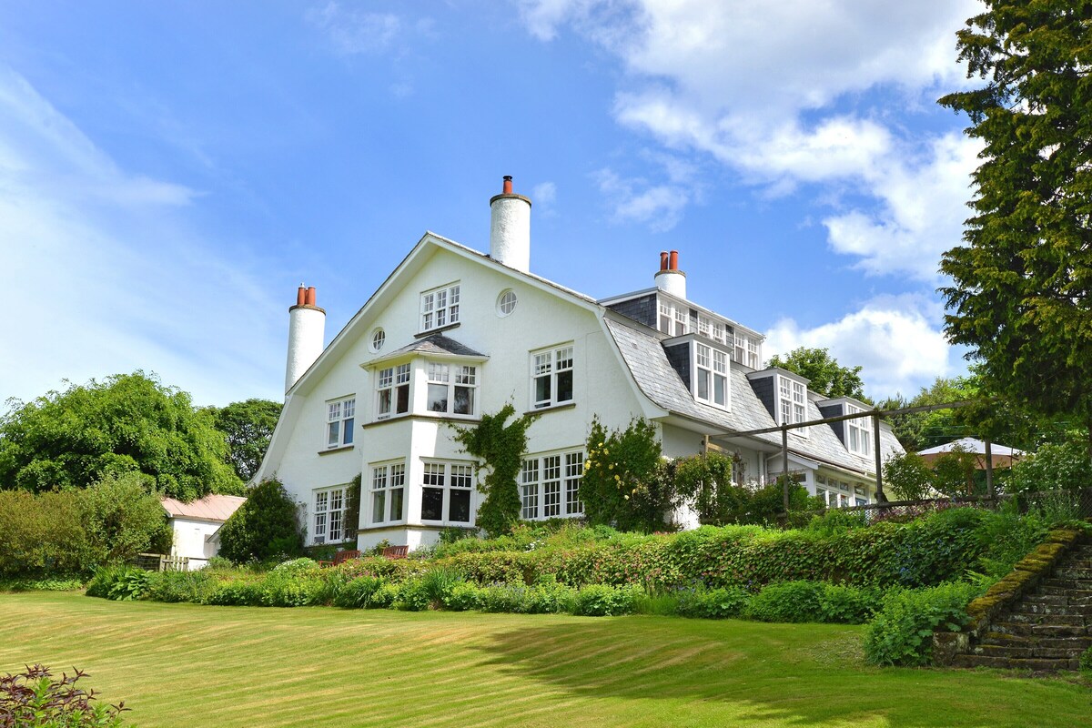A large, white, two-story house with multiple chimneys, surrounded by lush greenery and a well-maintained lawn.