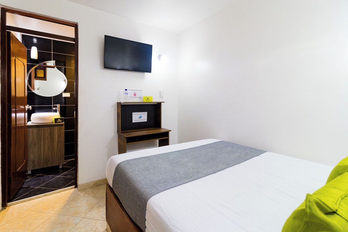 A hotel room with a bed, a wardrobe, a mirror, a television, and a small shelf with toiletries.