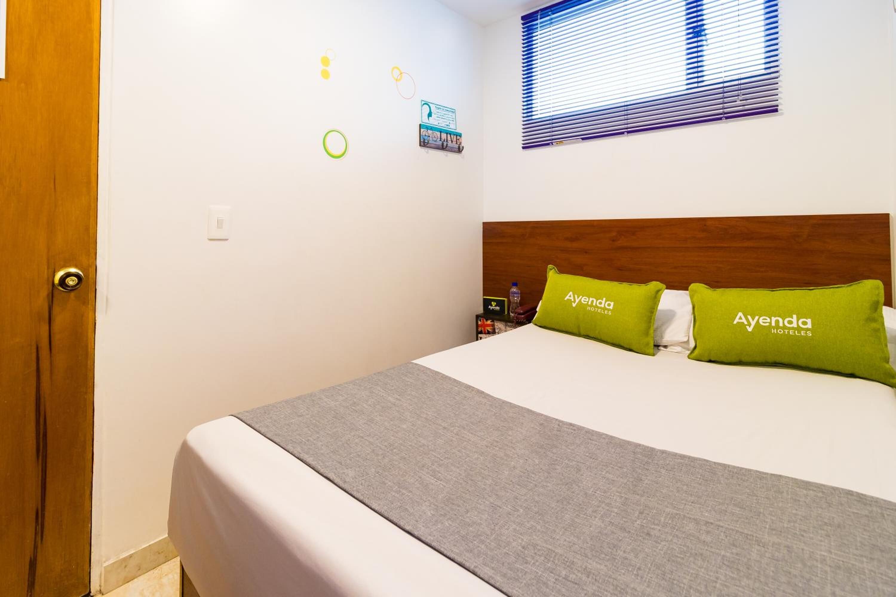 A hotel room with a bed, a wooden headboard, and a window with blinds.