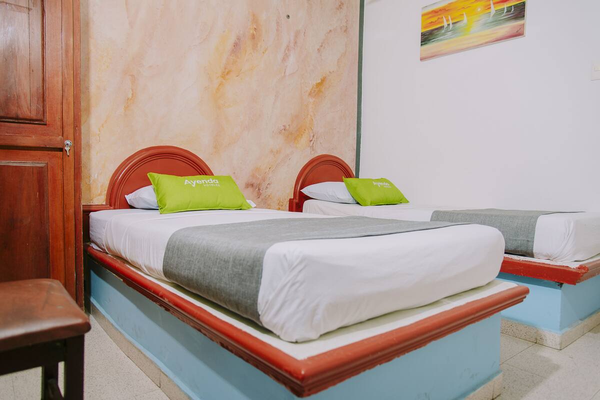 Ein Zimmer mit zwei Betten, jeweils mit einem hölzernen Kopfteil und weißer Bettwäsche. Auf den Betten liegen grüne Kissen, und links ist ein Holzsessel zu sehen.