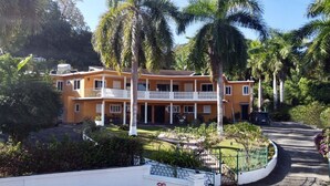 Um edifício de dois andares com varanda branca, rodeado por palmeiras e vegetação.
