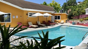 Uma área de piscina com espreguiçadeiras e guarda-sóis, rodeada por vegetação e um edifício amarelo.