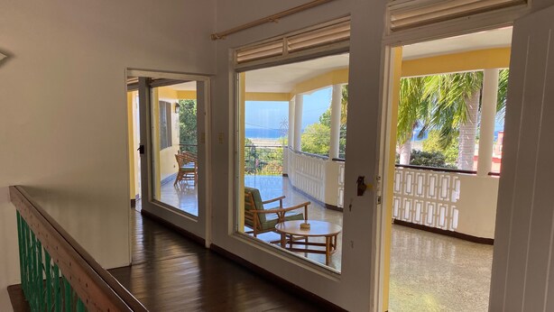 Um corredor com piso de madeira e corrimão verde, que leva a uma varanda com mobiliário externo e vista para o oceano.
