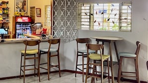 Um café com cadeiras de madeira e chão de azulejos, um balcão com refrigerador de bebidas e uma janela com vista para o verde.