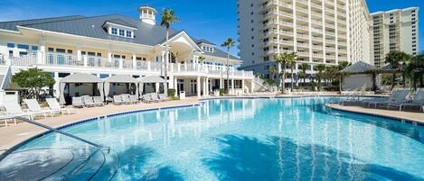 A large swimming pool with lounge chairs and umbrellas, surrounded by a hotel building with multiple balconies and palm trees.