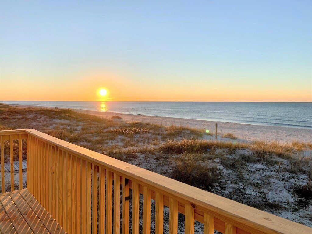 Una terraza de madera con vista a una playa y un atardecer al fondo.