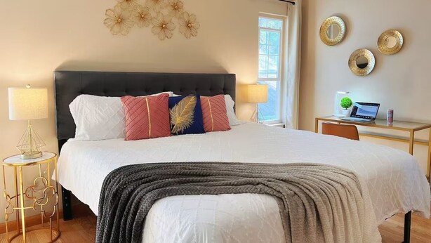 A bedroom with a bed, bedside lamps, a desk with a laptop, decorative wall art, and a window.